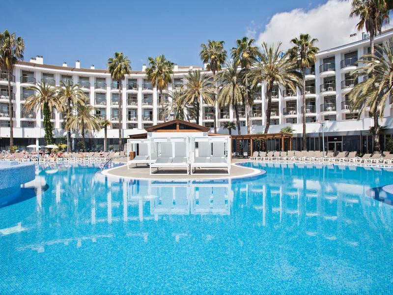 Gran piscina de hotel con palmeras y edificio blanco de varios pisos al fondo bajo cielo azul.