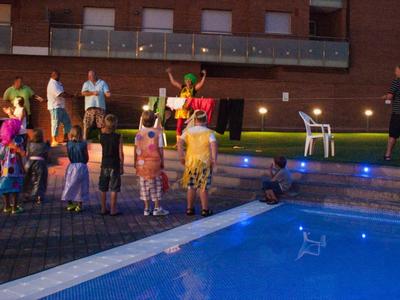 Gruppe bunter Tänzer in Kostümen steht neben blau beleuchtetem Pool vor rotem Backsteingebäude.