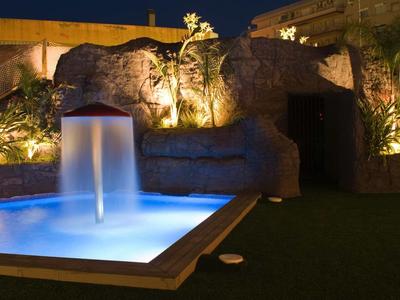 Beleuchteter Gartenbrunnen mit blauem Wasser vor einer Felsenwand bei Nacht.