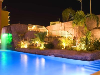 Nachtaufnahme eines beleuchteten Pools mit Wasserfall und warmen Gartenlichtern.