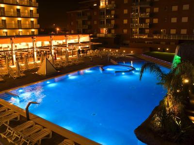 Beleuchteter Swimmingpool bei Nacht mit Liegestühlen und angrenzendem Restaurant in Mehrfamilienhaus.