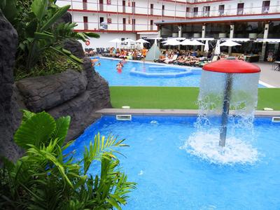 Pool with water feature and lounge area in front of a hotel building with balconies.
