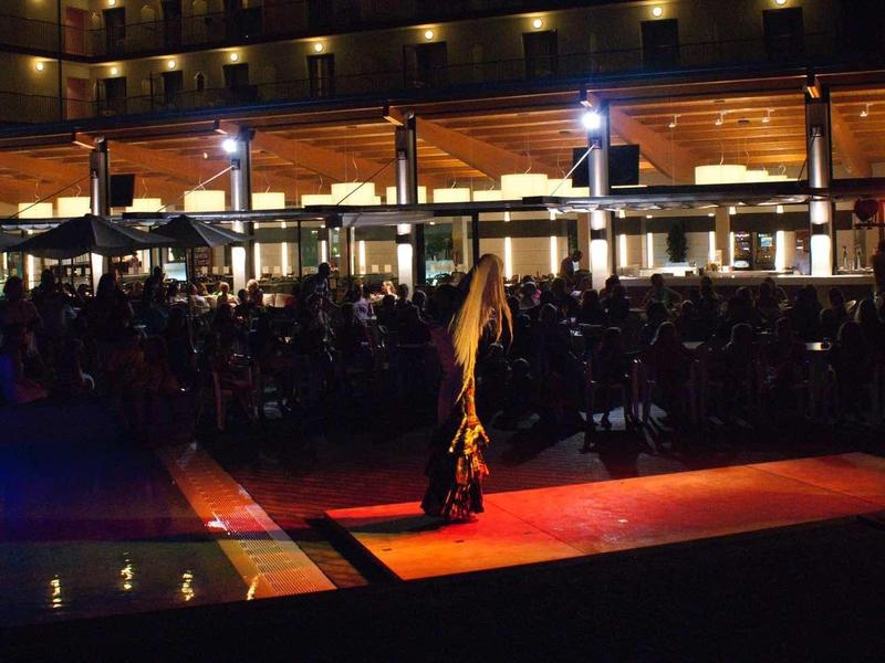 Dunkle Szene mit Lagerfeuer vor beleuchtetem Gebäude und Personen in der Dämmerung.