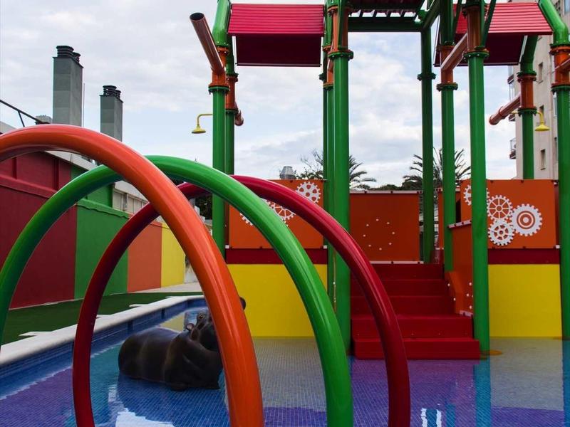 Bunte Wasserspielplatzstruktur mit Rutschen und Bögen, umgeben von einem Wasserbecken.