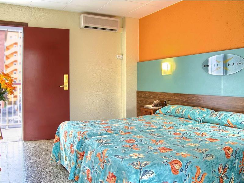 Schlafzimmer mit Doppelbett, buntem Bettbezug, offenem Fenster und Holzmöbeln in warmen Farben.