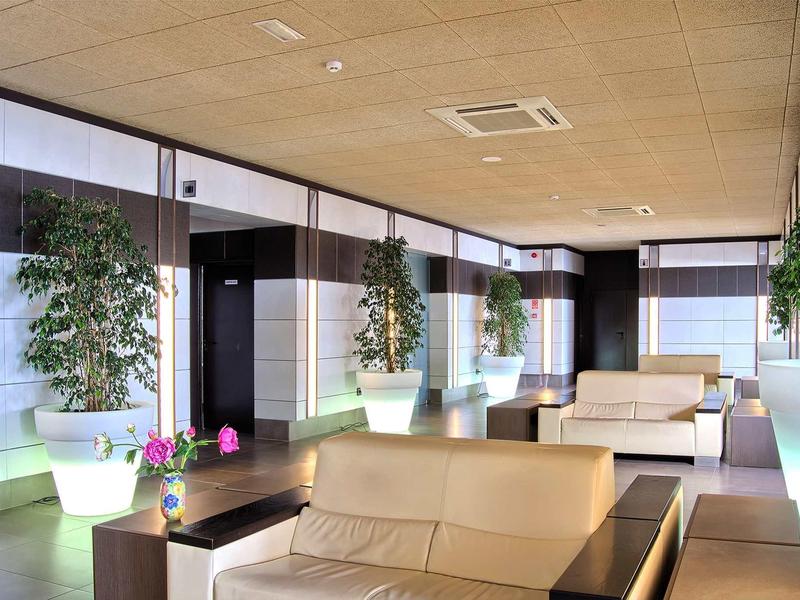 Moderne Lounge mit beigen Sofas, großen Pflanzen in beleuchteten Töpfen und Holzdecke.