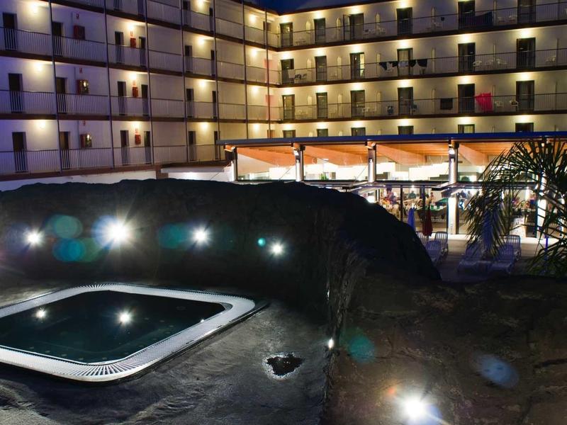 Hotel mit mehrstöckigem Gebäude, beleuchteten Balkonen und beleuchtetem Whirlpool im Vordergrund bei Nacht.