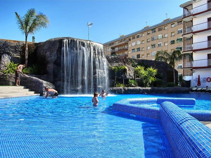 Schwimmbad mit Wasserfall, blauer Wasseroberfläche und umliegenden Gebäuden bei Sonnenschein.