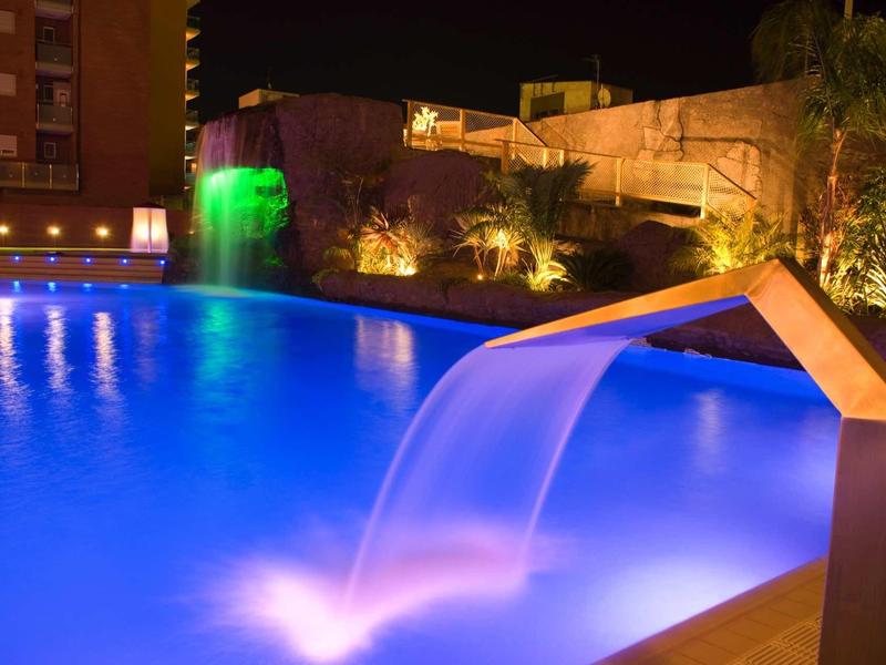 Beleuchteter Pool bei Nacht mit Wasserfall und moderner Architektur im Hintergrund.