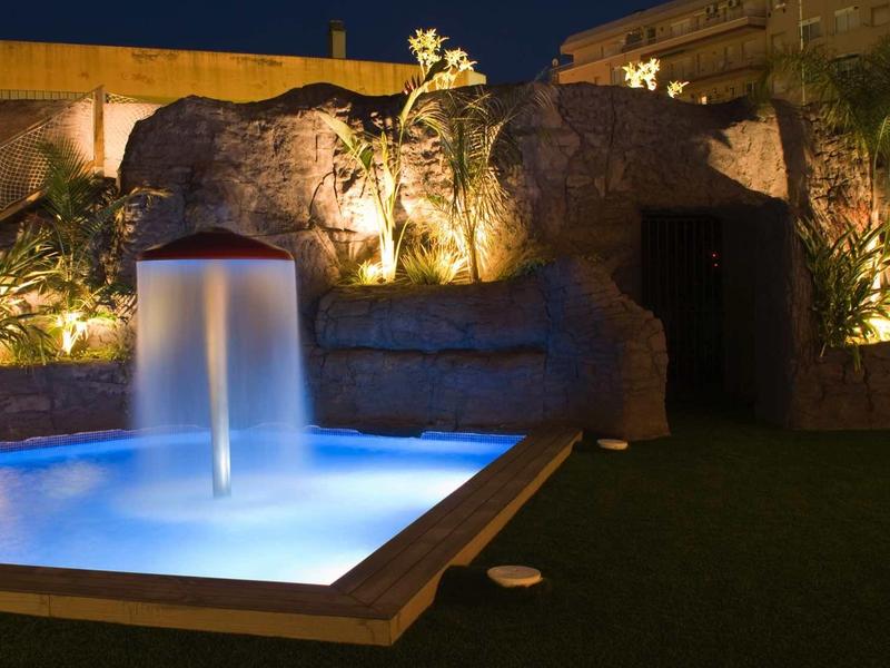 Beleuchteter Gartenbrunnen mit blauem Wasser vor einer Felsenwand bei Nacht.