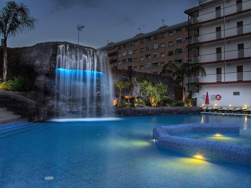 Beleuchteter Pool mit Wasserfall, Palmen und großen Gebäuden im Hintergrund bei Dämmerung.