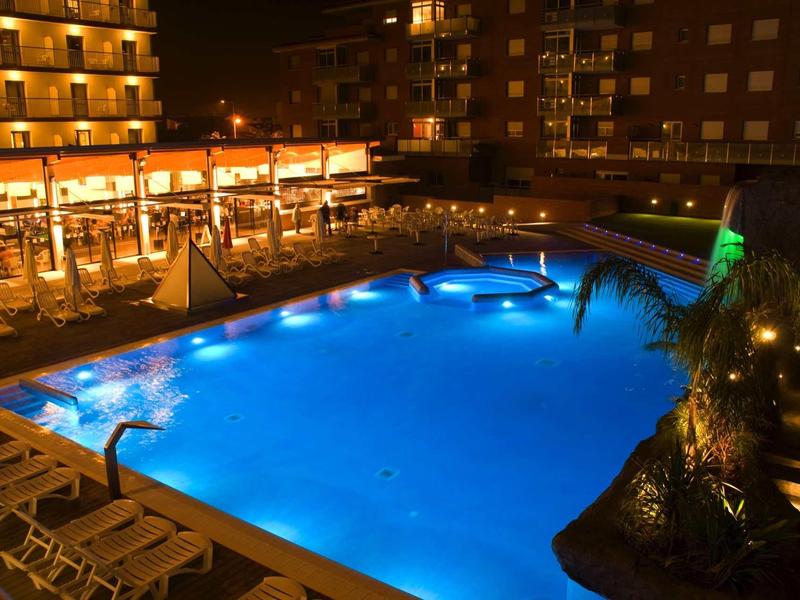 Beleuchteter Swimmingpool bei Nacht mit Liegestühlen und angrenzendem Restaurant in Mehrfamilienhaus.