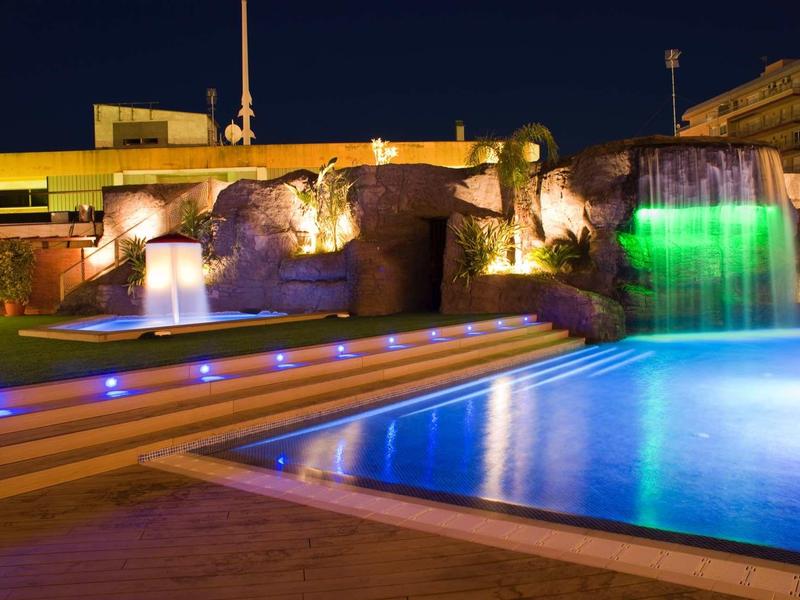 Nächtlicher Pool mit beleuchtetem Wasserfall und bunten Lichtern am Rand und Treppen.