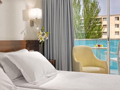 Hell eingerichtetes Schlafzimmer mit weißem Bett, gelbem Sessel und Blick auf Balkon und Bäume.