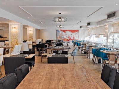 Heller, moderner Speisesaal mit Holztischen, schwarzen und blauen Stühlen sowie großen Fenstern.