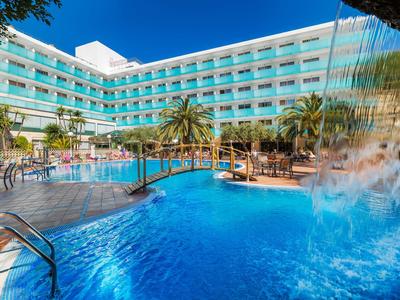 Großer Hotelpool mit Wasserfall, Holzbrücke, Palmen und blauem Himmel im Hintergrund.