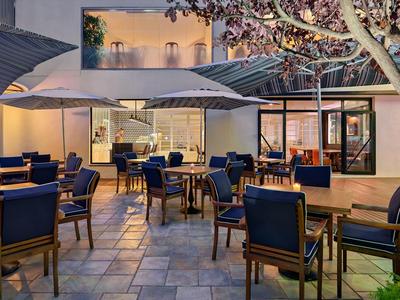 Moderner Außenbereich mit Holztischen, blauen Stühlen und großen Sonnenschirmen bei Abendlicht.