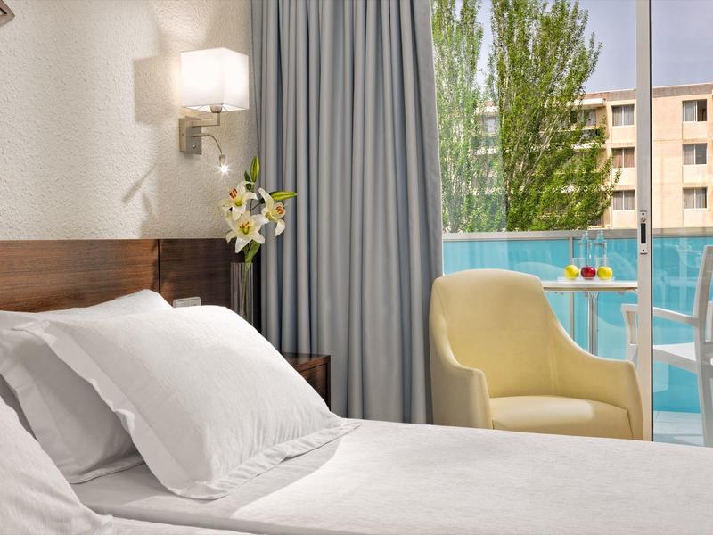 Hell eingerichtetes Schlafzimmer mit weißem Bett, gelbem Sessel und Blick auf Balkon und Bäume.