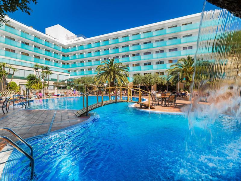 Großer Hotelpool mit Wasserfall, Holzbrücke, Palmen und blauem Himmel im Hintergrund.