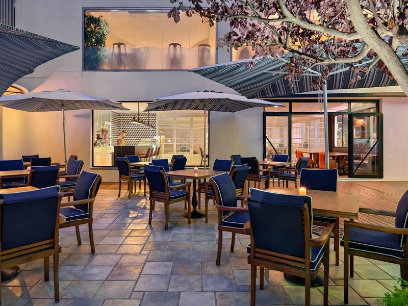 Moderner Außenbereich mit Holztischen, blauen Stühlen und großen Sonnenschirmen bei Abendlicht.