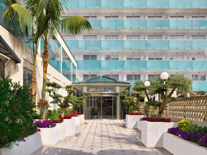 Eingang mit Glasfassade, Palmen und bunten Blumen in weißen Blumenkästen vor modernem Gebäude.