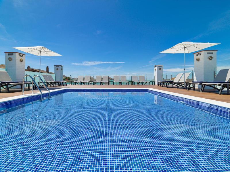 Schwimmbecken mit blauer Kachelfassade, Sonnenliegen und weißen Sonnenschirmen unter klarem Himmel.