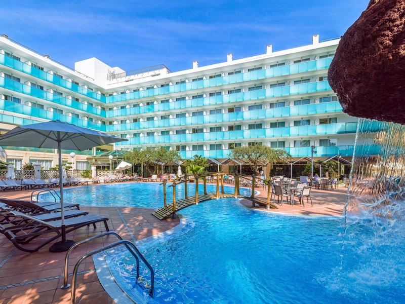Hotel mit großem Pool, Poolliegen und Wasserfall unter blauem Himmel.