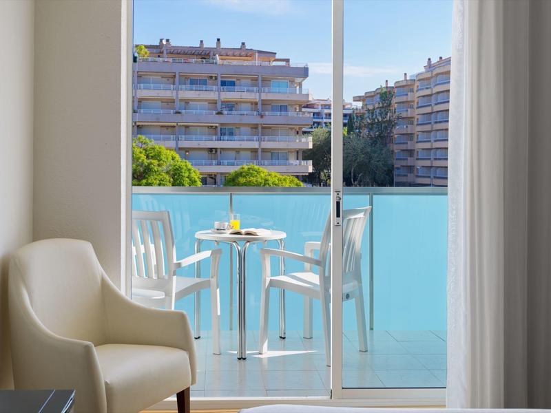 Balkon mit weißem Tisch und Stühlen, bequemer Sessel innen, Blick auf Wohnhäuser und blauen Himmel.