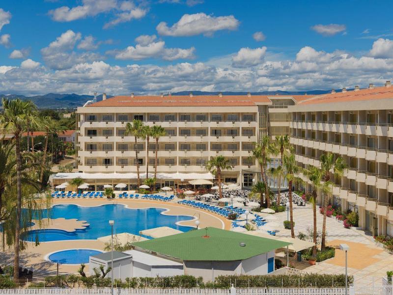 Duży hotel z basenem na zewnątrz, palmami i miejscami do siedzenia pod niebem z chmurami.