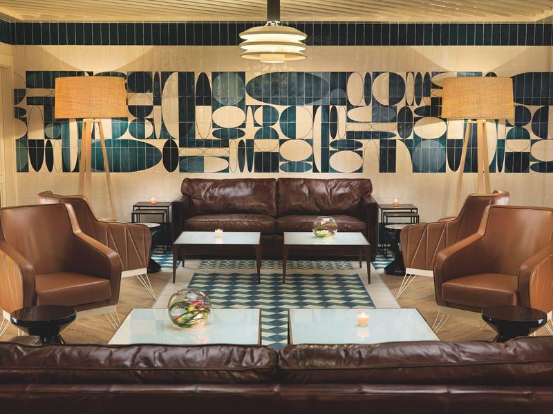 Przytulny salon hotelowy z brązowymi skórzanymi sofami, fotelami, szklanymi stolikami kawowymi i abstrakcyjną sztuką na ścianie.