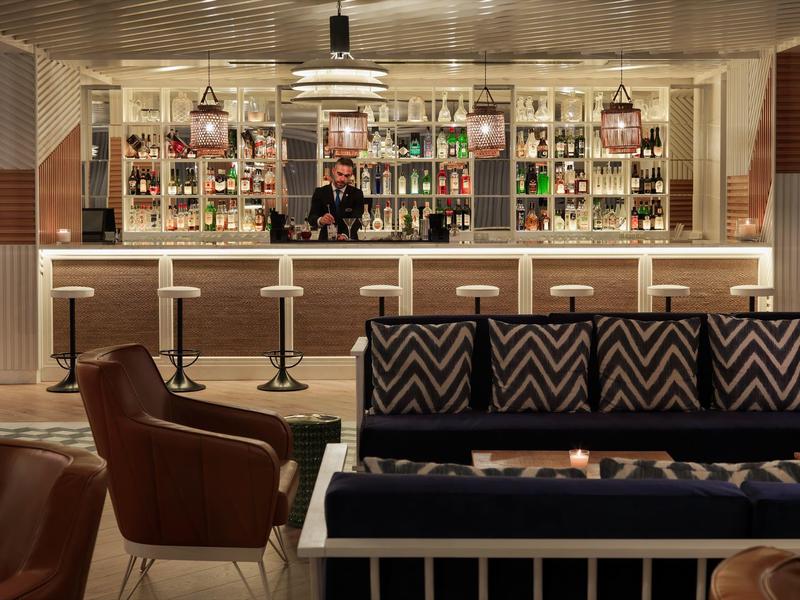 Nowoczesny bar hotelowy z hokerami, barmanem, brązowym fotelem i wzorzystą sofą.