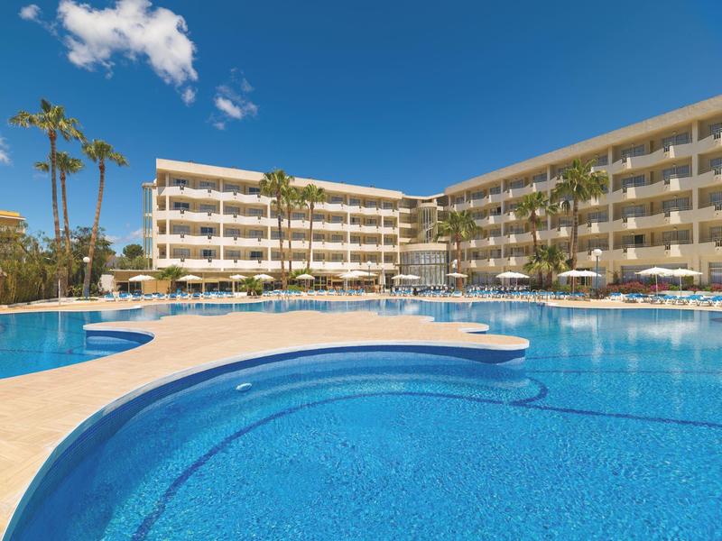 Duży hotel z wieloma balkonami i czystym, niebieskim basenem na pierwszym planie pod słonecznym niebem