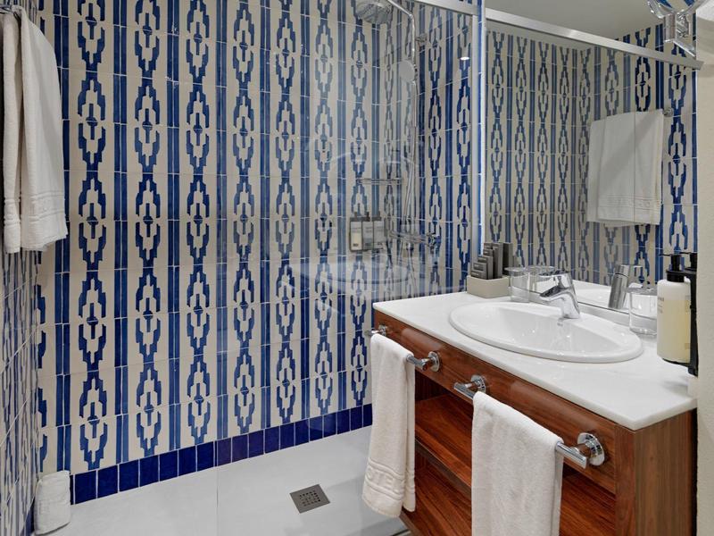 Nowoczesna łazienka z przeszklonym prysznicem, wzorzystymi niebieskimi płytkami, drewnianą szafką i białymi ręcznikami.