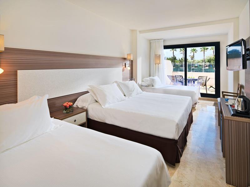 Pokój hotelowy z trzema łóżkami, białą pościelą, drewnianymi zagłówkami i widokiem na balkon.