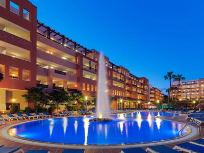 Basen hotelowy z oświetloną fontanną o zmierzchu, otoczony oświetlonymi balkonami i palmami.