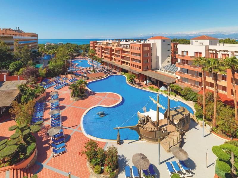 Hotel z dużym nieregularnym basenem, otoczony leżakami i palmami pod jasnym niebem.