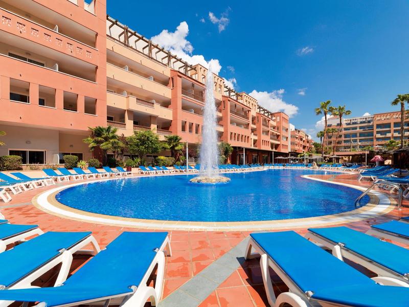 Basen hotelowy z leżakami i fontanną pod jasnym niebem.