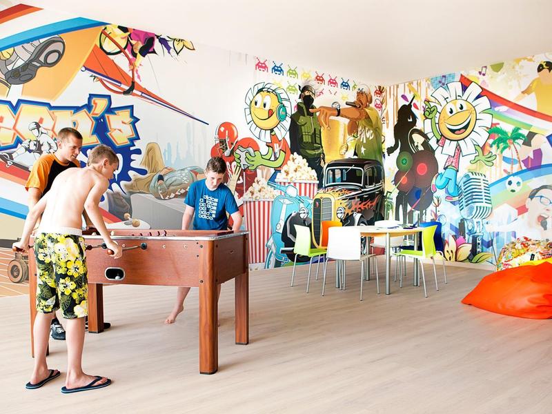 Jasny pokój zabaw z kolorowymi ścianami graffiti, dzieci grające w piłkarzyki i pufy.