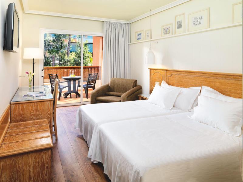 Pokój hotelowy z dwoma łóżkami, drewnianymi meblami, fotelem i balkonem z miejscem do siedzenia.