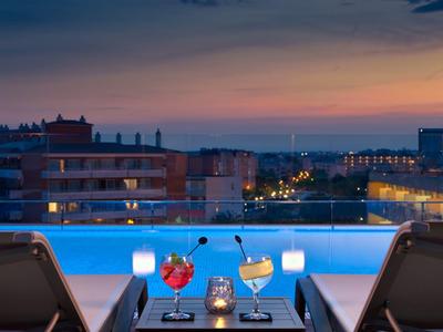 Terrasse avec piscine, vue sur la ville au crépuscule, deux transats et boissons sur table.