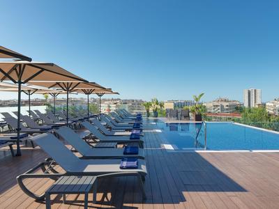 Moderner Hotelpool mit Liegestühlen und Sonnenschirmen auf einem Holzdeck unter blauem Himmel.