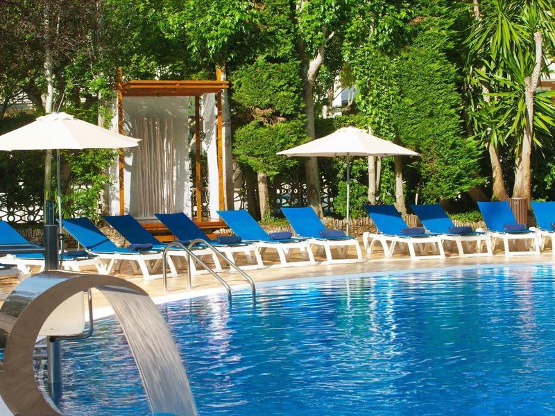 Chaises longues bleues et parasols blancs près d'une piscine bordée d'arbres verts.