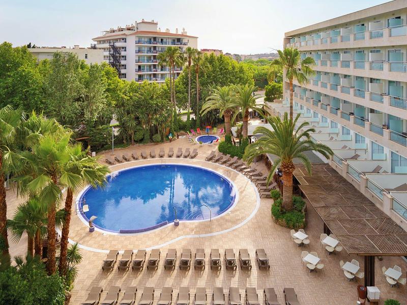 Piscine ovale entourée de transats près d'un hôtel et de palmiers.