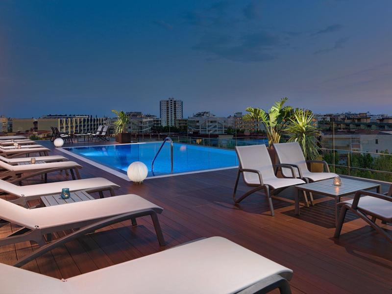 Piscine extérieure sur une terrasse d'hôtel avec chaises longues et vue sur la ville au crépuscule.