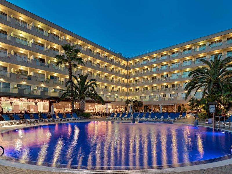 Hôtel moderne avec grande piscine éclairée et chaises longues sous ciel bleu au crépuscule.