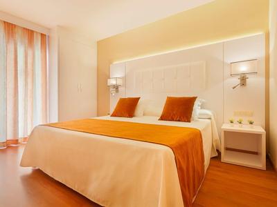 Světle zařízený hotelový pokoj s manželskou postelí a oranžovým přehozem a polštáři