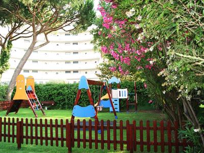 Dětské hřiště se skluzavkou a prolézačkou, obklopené stromy a keři poblíž budovy.