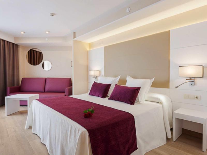 Moderní hotelový pokoj s manželskou postelí, červenými polštáři a pohovkou před hnědými závěsy.