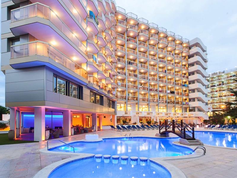 Moderní hotel se osvětlenými balkony a velkým venkovním bazénem za soumraku.