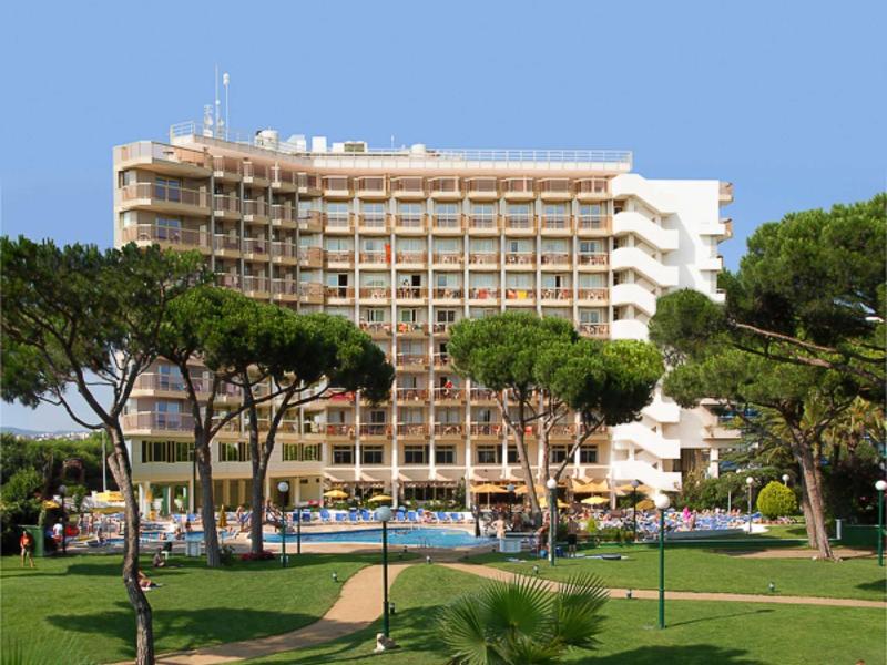 Vysoký hotel s balkónovými pokoji obklopený stromy a zelenými trávníky pod modrou oblohou.